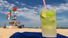 Um copo de caipirinha em area de praia, com pessoas e barracas ao fundo