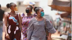 Women wear face masks with kente and wax print designs as they walk through a market.