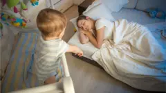 A baby in a cot looks at a mother asleep on a mattress close by