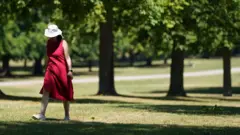 A woman in a red dress and sun hat walks by some trees
