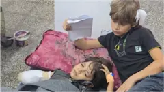 一名手臂纏著繃帶的女孩躺在卡邁勒阿德萬醫院的地板上，一名男孩陪伴在她身邊。