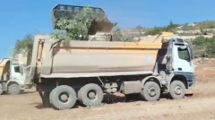 Zeytin ağac yüklü bir kamyon toprak bir arazide, arkasından bir başkası daha görünüyor. 