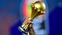 Le trophée de la Coupe d'Afrique des Nations est brandi par la main droite d'un homme, sur un arrière-plan flou.