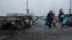 Mulher e criança segurando sacolas passando em frente a carros incendiados