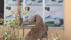 یوه سړي کمیس پرتوګ اغوستی، زړې څوکۍ باندې ناست دی، زوړ میز باندې یې خپل سر ایښی 