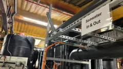 Tubulações dentro da instalação em Weymouth; uma placa diz: 'Água do Mar Entra e Sai'