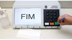 Uma mão pressiona o botão confirma de uma urna eletrônica com uma caneta