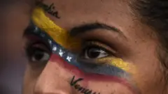 Imagen muy cerrada de los ojos de una mujer con la bandera colombiana y venezolana pintada en el rostro y las palabras "Colombia" y "Venezuela".