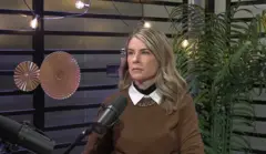 Rita Lobo é uma mulher branca e loira. Ela está sentada em um estúdio para uma entrevista. Na frente dela há um microfone. Ela usa um suéter marrom.