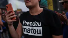 Uma pessoa vestindo uma camiseta preta com os dizeres "prendeu matou".