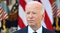 US President Joe Biden looks into the distance. He wears a pinstriped suit and has American flags behind him.