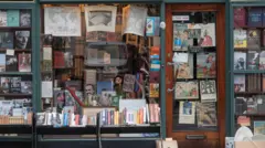 通り沿いの書店のウィンドウや入口にポスターが貼られ、ディスプレイにさまざまな本が並べられている。ウィンドウ越しに店内の書棚も見える。店の外にも低い棚が置かれ、本が並べられている