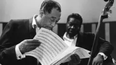 Djuk Elington drži notnu svesku iza njega drugi muzičar 