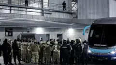 Migrantes deixando o bloco 8 do Cecot em direção a um ônibus, cercados por guardas