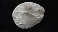 Um pedaço arredondado de sílex que foi cuidadosamente trabalhado para criar uma faca. É branco, com algumas marcas marrons e cinza, e é colocado sobre um fundo preto. As áreas onde pedaços do sílex foram arrancados com outra pedra são claramente visíveis. Não é um círculo perfeito, mas seus lados são arredondados.