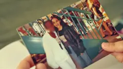 Uma fotografia impressa é segurada por duas mãos. A imagem mostra Isabel na adolescência, sorrindo e posando ao lado de uma amiga, cujo rosto foi desfocado. As duas aparecem em frente a grades metálicas azul-escuras, em um local público movimentado. Isabel veste uma jaqueta escura e calça jeans, enquanto a outra pessoa está vestida de branco.