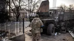 Umusirikare wa Ukraine ahagaze iruhande rw'imodoka y'ikamyo y'ingabo z'Uburusiya, mu mujyi wa Trostianets mu majyaruguru ashyira uburasirazuba