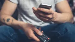 Hombre con un mando a distancia en una mano y un smartphone en la otra.