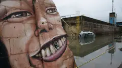 Um grande mural de rosto humano ocupa um muro de concreto, com destaque para o seu sorriso. Ao lado, uma área está alagada e um carro aparece parcialmente submerso na água. O ambiente é cinzento.