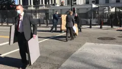 Homens usando máscaras e vestidos com ternos carregam painéis e caixas de papelão na rua diante da Casa Branca, durante o dia