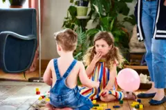 Uma menina brincando com um menino de montar blocos de madeira no chão de uma casa — ela está em frente a ele tapando a boca com a mão