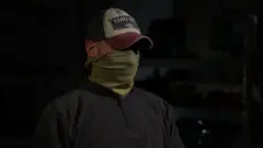 Ruslan, Forças de Operações Especiais, com um chapéu e um gorro cobrindo o rosto e a identidade em um quarto escuro, vestindo um moletom preto.