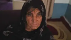 Sabbah, mãe de Fazel e Naik, aparece de véu e olhando para a câmera em foto tirada dentro de ambiente interno