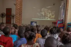 Les pensionnaires de l'orphelinat, de dos, regardent un film projeté sur grand écran dans un hall.