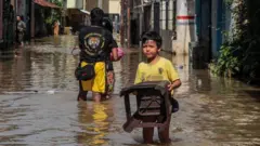 A boy carrying a chair walks through a submerged road in Indonesia
