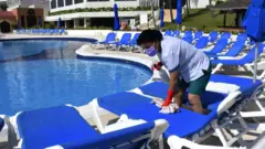 Trabalhadora com máscara limpa espreguiçadeiras no entorno de piscina de resort