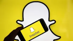 The Snapchat ghost logo is seen as a background with a phone and the app held up in front of it