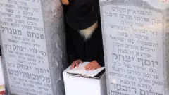 Rabino agachado diante de livro sagrado, entre dois túmulos