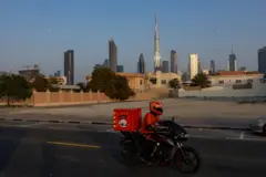 यूएईमा सामान डेलिभरी गर्न मोटरसाइकल चढेका एक व्यक्ति, पृष्ठभूमिमा अग्ला भवनहरू छन्