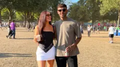 Lauren Macpherson aparece ao lado do namorado, Zak. Os dois estão em um festival. Ela tem cabelos longos e escuros e usa uma saia branca e um top preto tipo bandana, enquanto ele veste uma camiseta bege e jeans pretos