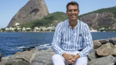 Tim Vickery sentado em rochas com o o mar e os morros do Rio de Janeiro ao fundo