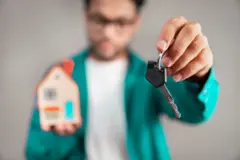 Young man holding house model and key in hand. He is standing against an isolated gray background.