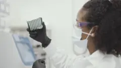 Mulher de jaleco olha para amostras de vacina em ambiente de laboratório