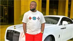 Hushpupi in front of a luxurious car