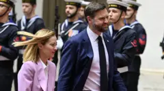 Giorgia Meloni, que tem cabelos loiros na altura dos ombros e usa um terno lilás, passa por soldados italianos com o presidente dos EUA, JD Vance, que tem cabelos castanhos curtos e penteados e usa um terno azul-marinho com camisa branca e gravata preta, no Palazzo Chigi, em Roma.