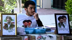 Um vendedor aguarda clientes enquanto vende porta-retratos do falecido Líder Supremo do Irã, o aiatolá Ali Khamenei, ao lado de seu filho e atual Líder Supremo, Mojtaba Khamenei, do lado de fora de uma mesquita xiita em Islamabad, em 5 de abril de 2026
