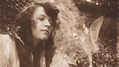 Un hada parada en la rama de un árbol, ofreciéndole a Elsie un pequeño ramo de flores.