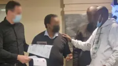 ጆን ሐብቴ [ከመሃል] ከፖሊሶች ጋር