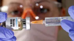 Photograph of a woman wearing gloves and PPE putting a syringe into a bottle marked Covid-19