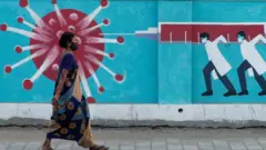 A woman wearing a mask walking past graffiti of three doctors injecting an virus