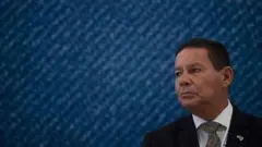 Mourão olhando para o lado, com olhar sério, em frente a painel azul