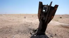 Um fragmento de um míssil balístico jaz parcialmente soterrado no deserto, tendo ao fundo um céu azul limpo e uma figura solitária fora de foco