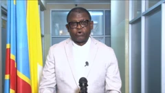 Musenyeri Donatien Nshole, umunyamabanga mukuru wa CENCO, arimo gusoma ubutumwa bw'iyo nama nkuru y'abasenyeri muri Repubulika ya Demokarasi ya Congo. Ahagaze imbere y'indangururamajwi, yambaye ikoti rya khaki rijya kuba umweru ricometseho indangururamajwi ntoya, n'ishati y'umweru iriho ikora rigufi rimenyerewe ku bihayimana. Iruhande rwe hari ibendera rya Congo. 