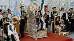 Coroação de Mohamed Reza Pahlavi. Ao lado esquerdo dele está uma criança e do lado direito, sua esposa, sentada em um cadeira e usando uma coroa