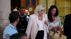 Marine Le Pen, una mujer rubia con chaqueta rosada, camisa blanca y falda negra, sale del Tribunal de Apelaciones en Francia, acompañada de una asistente y seguida de un guardia con chaleco antibalas.