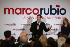 Marco Rubio falando no microfone. Ao fundo, há um painel com o nome dele